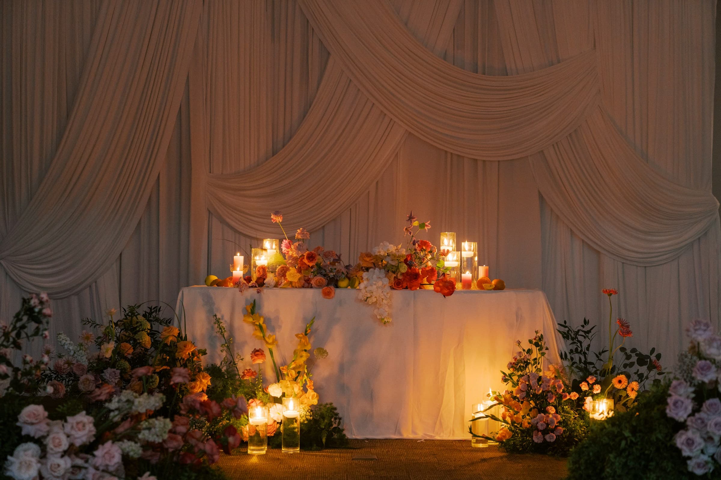 Wedding floral setup on table covered in white linen, with candles and floor covered in more flowers and white drapery